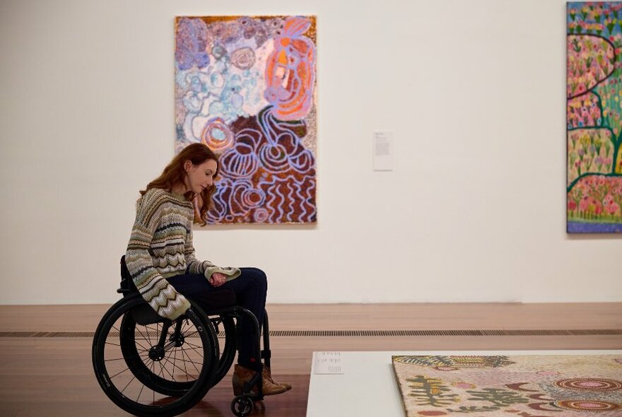 Person in a wheelchair inside an art gallery looking at a large piece of Aboriginal art on the floor.