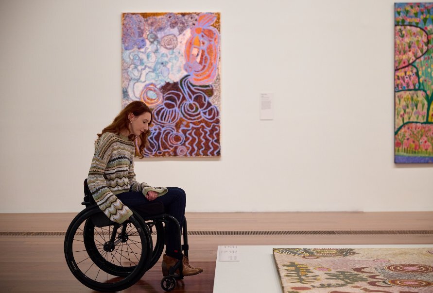 Person in a wheelchair inside an art gallery looking at a large piece of Aboriginal art on the floor.