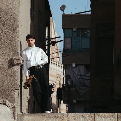 The musical artist Saint Levant, wearing dark trousers and a white shirt holding a saxophone by his side, leaning against a wall, with buildings in the shadows behind him.