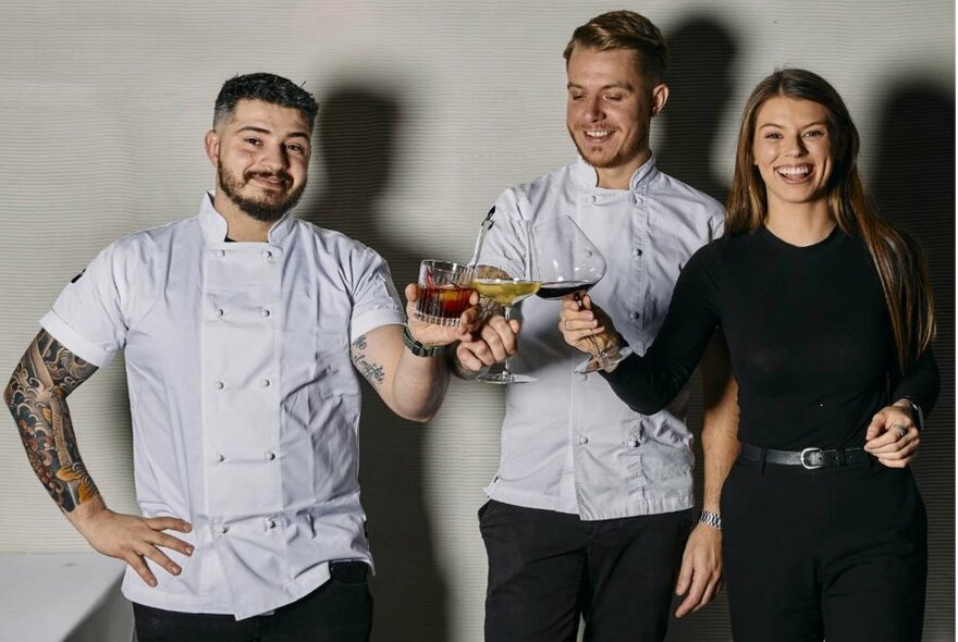 Two men in chef whites, and woman in black, all holding glasses and 'cheersing'.