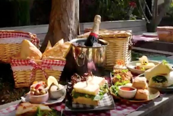 Picnic hamper ingredients spread out on a table, including a bottle of sparkling wine, cheese, bread and assorted nibbles.