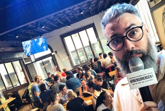 Trivia Host Ben Sorensen looking awkwardly at the camera with a microphone, a room full of people behind him playing trivia at tables in a pub.