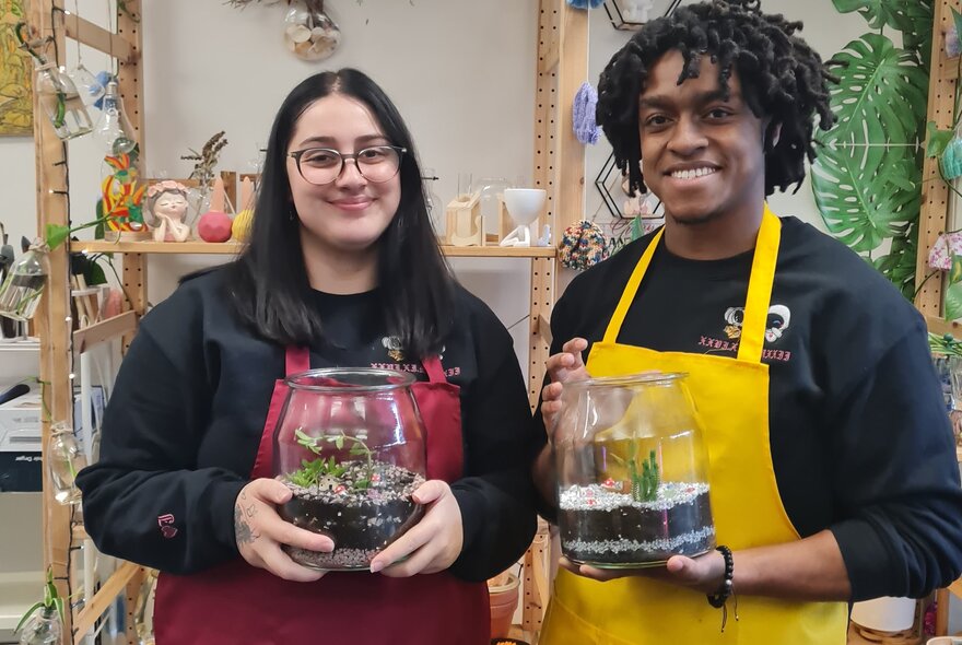Two participants in a studio workshop setting, holding glass-domed terrariums, smiling.
