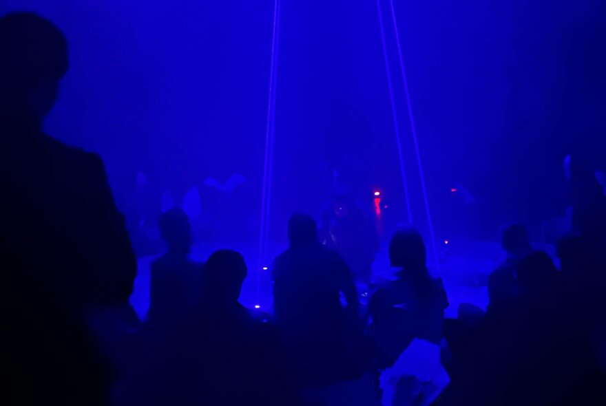 A dimly lit concert or performance venue bathed in intense blue light, silhouettes of the audience in the foreground.