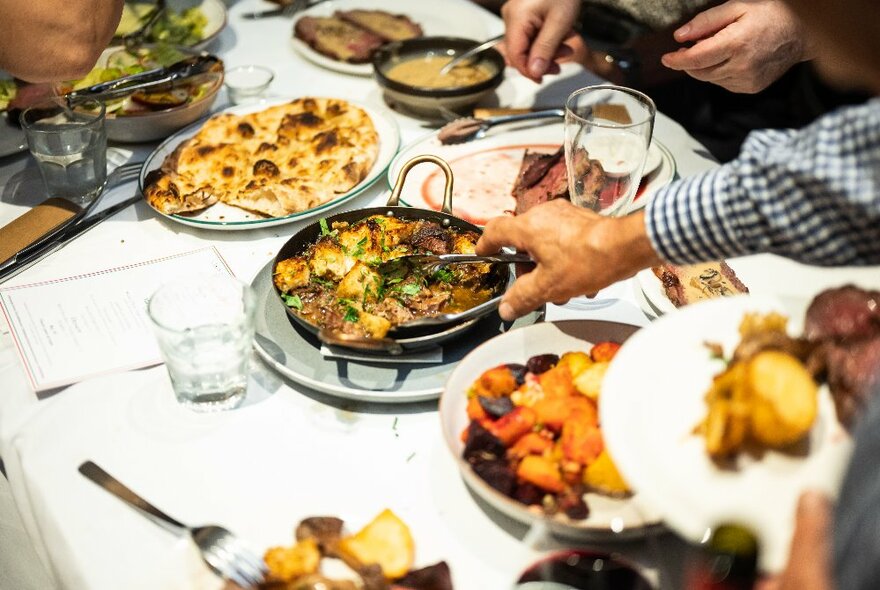 Plates of food on a white table, diners seated around it, with hands reaching in using tongs to select food.