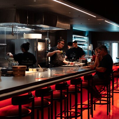 A dining couple seated on a high stool at the sleek bar area at Sachi restaurant, with a view into the  open kitchen space where several chefs are working. 