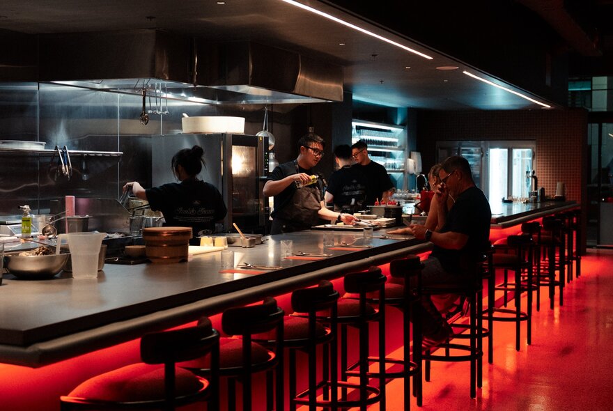 A dining couple seated on a high stool at the sleek bar area at Sachi restaurant, with a view into the  open kitchen space where several chefs are working. 