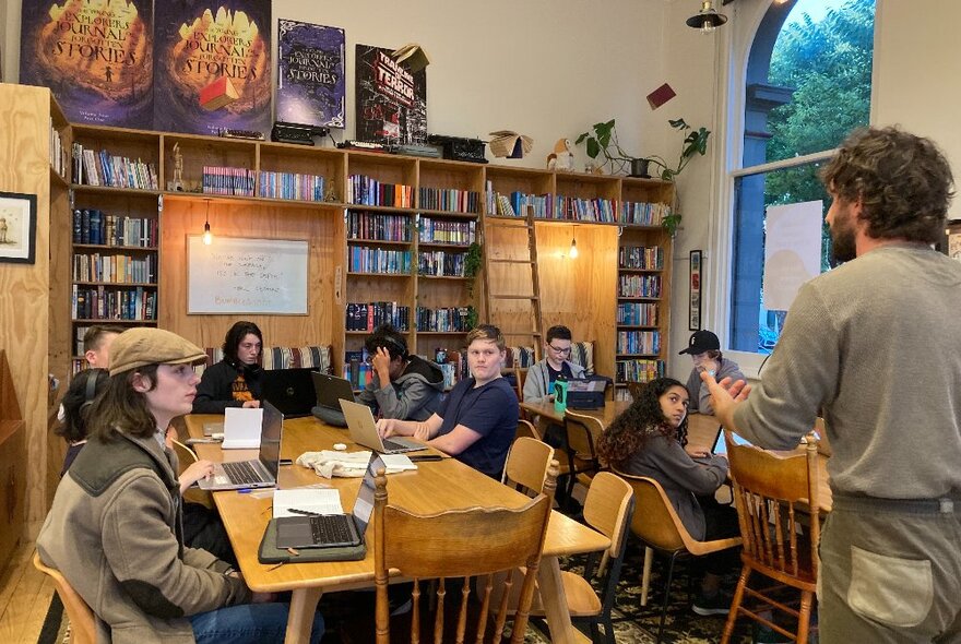 Teenagers sitting and working around two large tables in a room, some with laptops open in front of them, bookshelves in the background and a teacher standing in front of them talking to them.