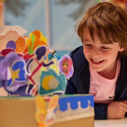 A boy smiling as he leans on a bench where handcrafted paper flowers are displayed standing up in a cardboard box.