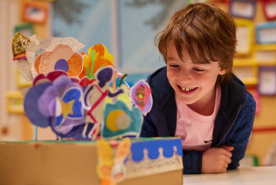 A boy smiling as he leans on a bench where handcrafted paper flowers are displayed standing up in a cardboard box.