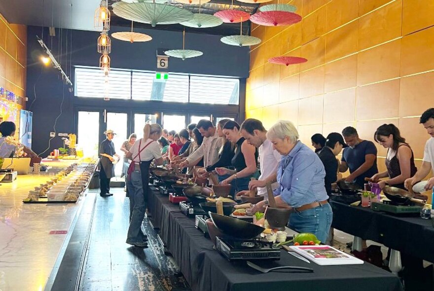 Cooking workshop participants standing behind two long tables set up with cooking stations, ingredients and utensils, inside a restaurant.