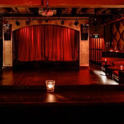 An intimate, dimly lit indoor stage venue with deep red velvet curtains drawn across a central archway, with booth seating around the walls surrounding the dancefloor.
