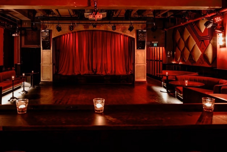 An intimate, dimly lit indoor stage venue with deep red velvet curtains drawn across a central archway, with booth seating around the walls surrounding the dancefloor.
