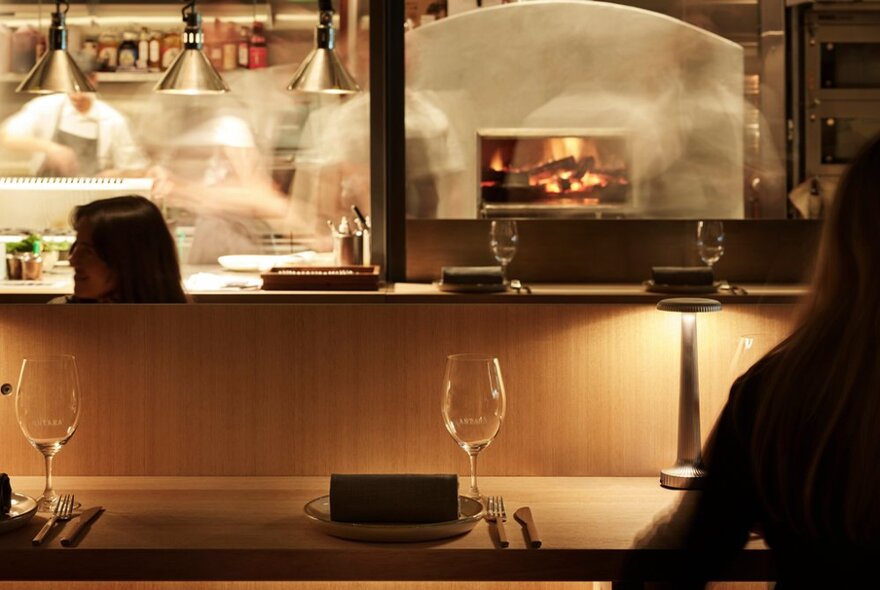 Bar area seating in front of the open kitchen of a restaurant, with chefs and a wood fired hearth in the background.