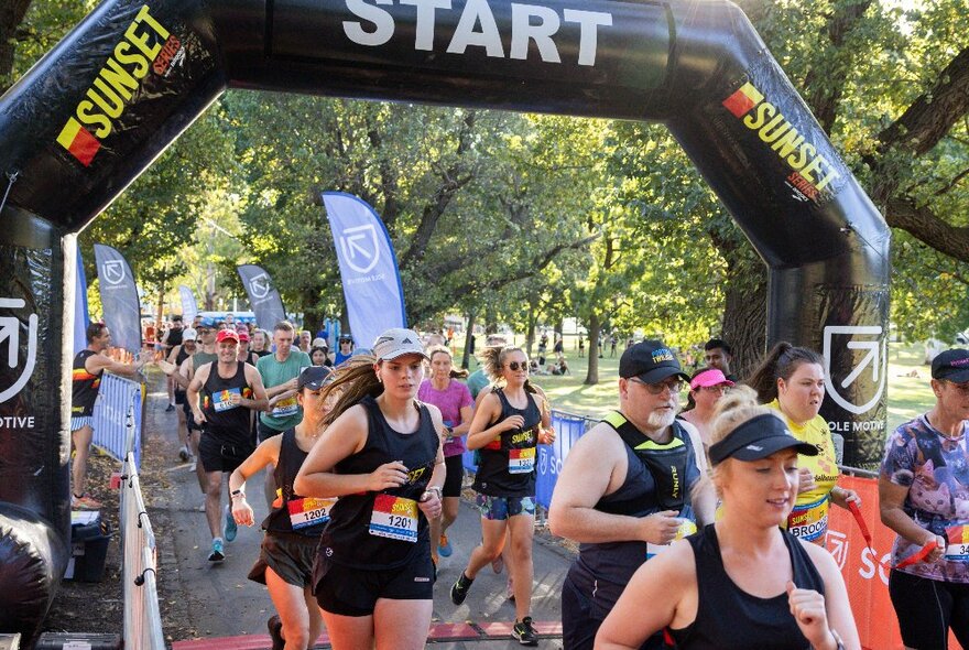 A large group of people running around Princes Park under an inflatable starting arch, large trees in the background.