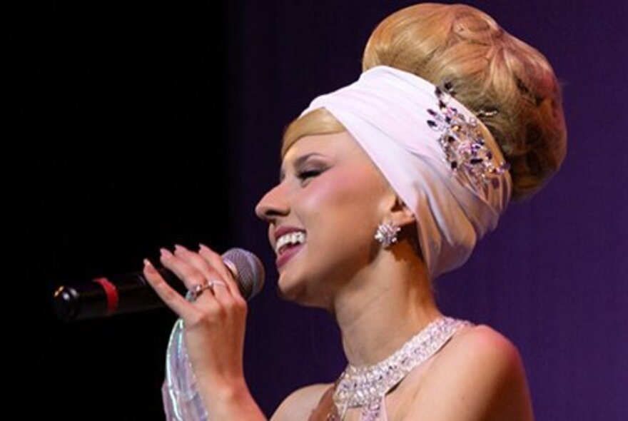 Singer Rachael Grace with her hair in a beehive hairdo and scarf, singing into a microphone she holds in her hands.