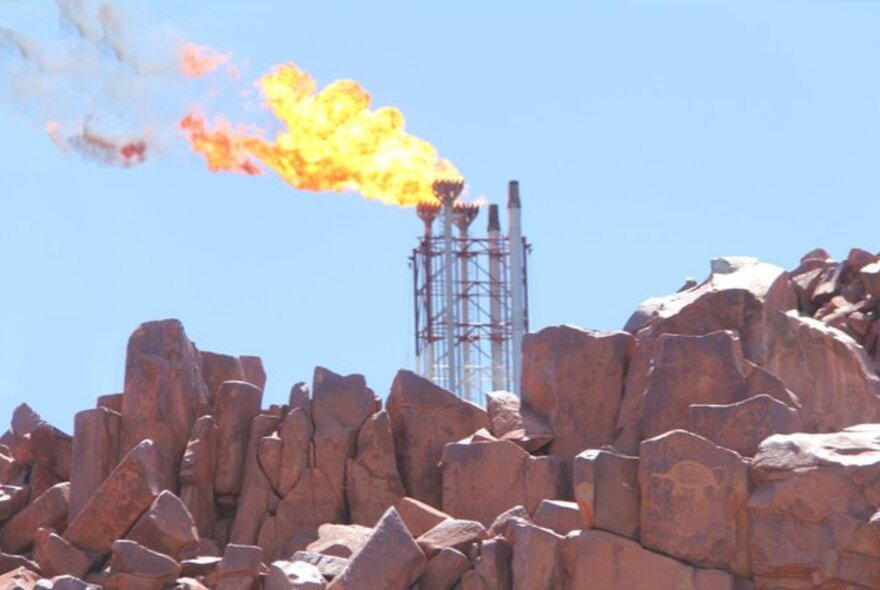 The flares from the top of an LPG tower against a blue sky, with an area of red rocks in the foreground.