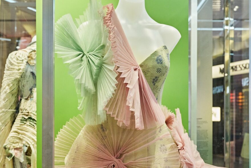 An avant-garde gown crafted with elaborate, fan-like accordion-pleated organza ruffles in contrasting pastel green and pink hues, displayed in a glass case in a public space.