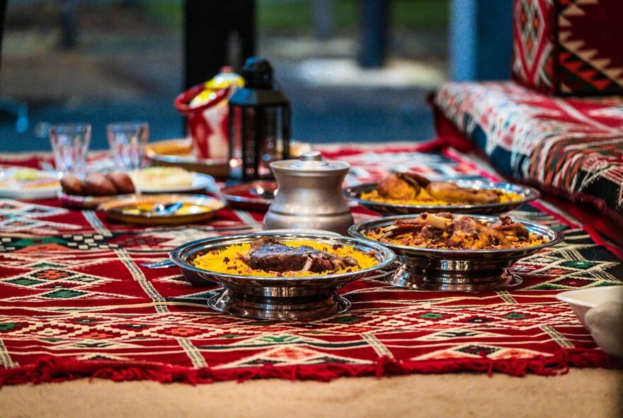 Silver platters of Yemeni food presented on red table covering on a low table with matching seating, including bowls of rice and meat, with glasses and table lantern.