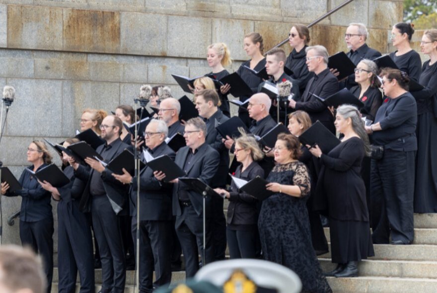 A chorus of singers all wearing black, standing on the steps of the Shrine of Remembrance performing a concert.