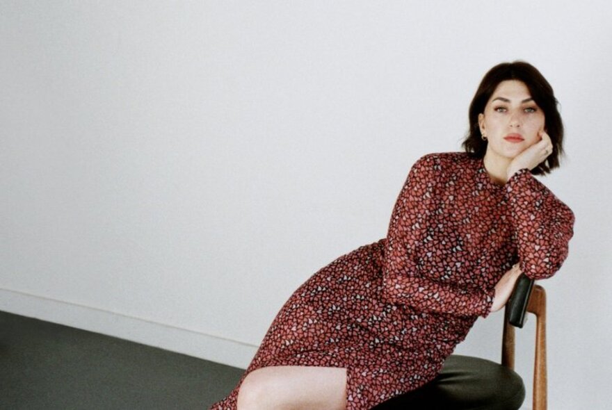 Jazz singer, Elissa Rodger, wearing a glittery, dark red evening dress and leaning on a small leather chair in front of a white wall.