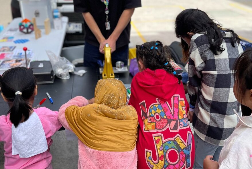 View from behind of four kids waiting at a craft activity table, outside.