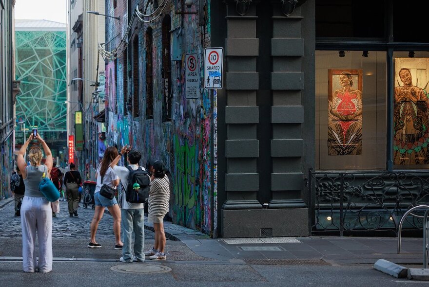 People gathered outside on a street corner looking down an alleyway with graffiti. 