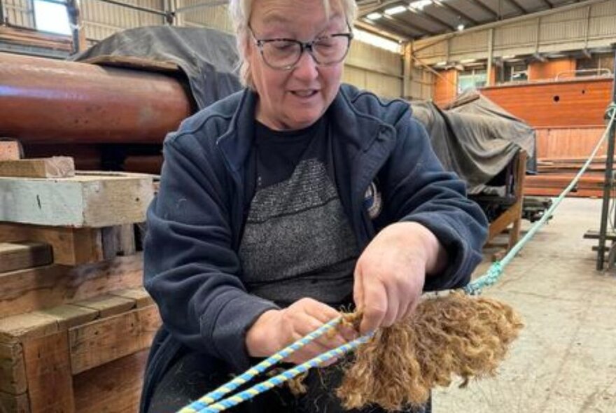 A woman tieing knots in ropes in a boatbuilding yard with equipment and wood.