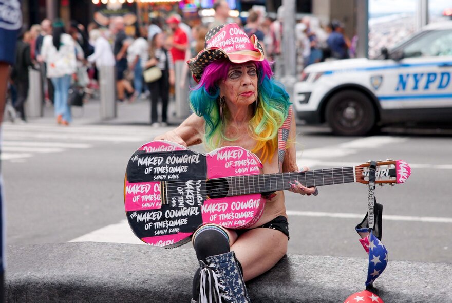 A person with brightly coloured long hair playing an acoustic guitar, outdoors on a New York city street, with cars and pedestrians in the background.