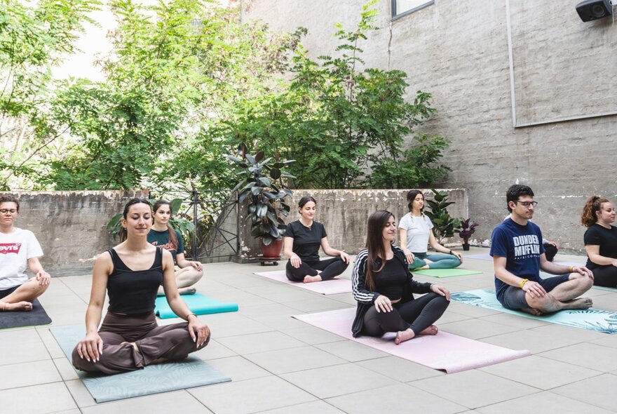People quietly sitting cross-legged on yoga mats in an outdoor courtyard, with green plants and dappled sunlight in the background.
