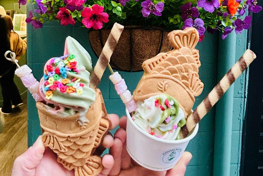 Two hands holding up waffles shaped like fish topped with soft-serve ice-cream, sprinkles and a biscuit stick, in front of a green wall. 