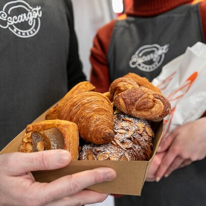 Two people wearing aprons, holding an array of flakey pastries and bread.
