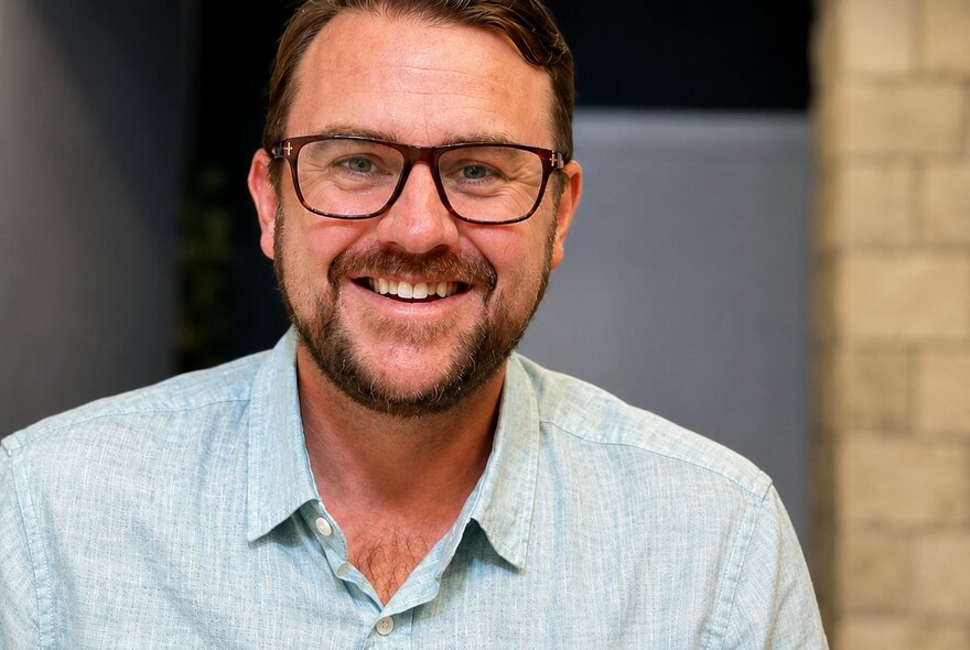 A man with glassing smiling openly and warmly in front of a blurred beige brick background.