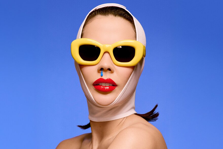 A person wearing large yellow sunglasses, bright red lipstick, a blue drip coming out from one of their nostrils, and a white compression bandage around their head and chin, against a solid blue background.