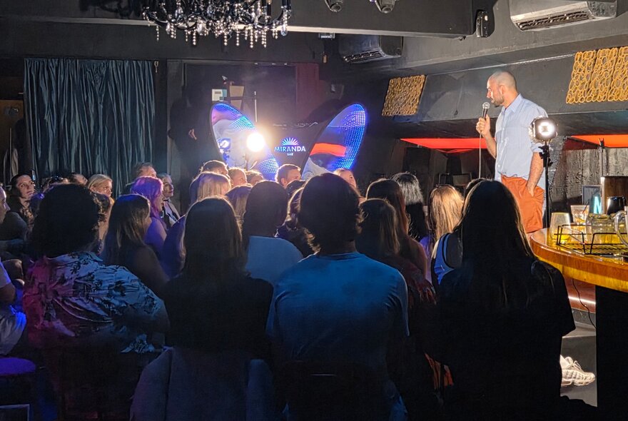 People seated in a dark bar venue watching a stand-up performer illuminated on stage, standing at a microphone.