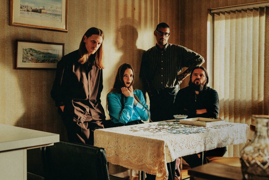The four members of the band Dry Cleaning in a dimly lit domestic interior, standing or seated around a kitchen table with a jigsaw puzzle in pieces in front of them.