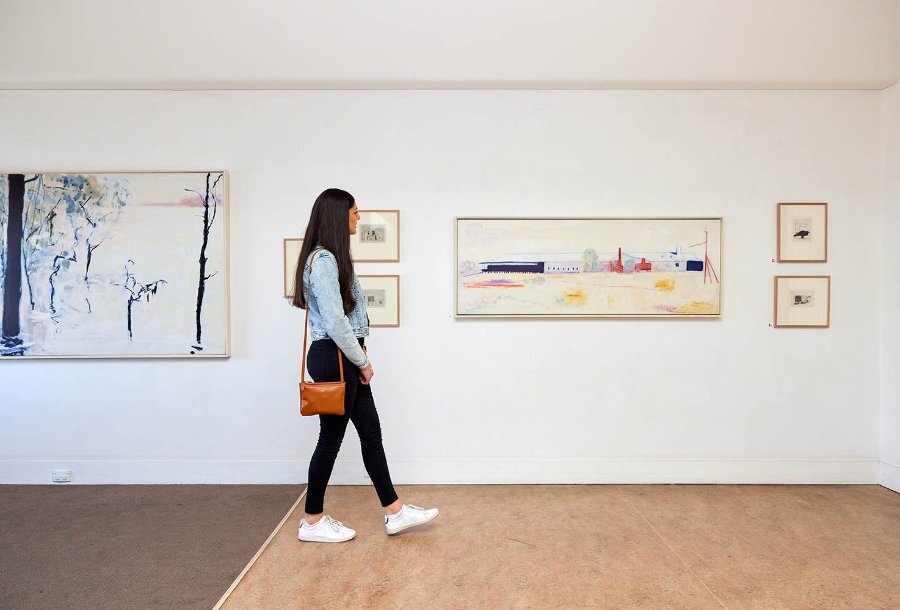 A woman walking through a gallery looking at paintings on the wall.