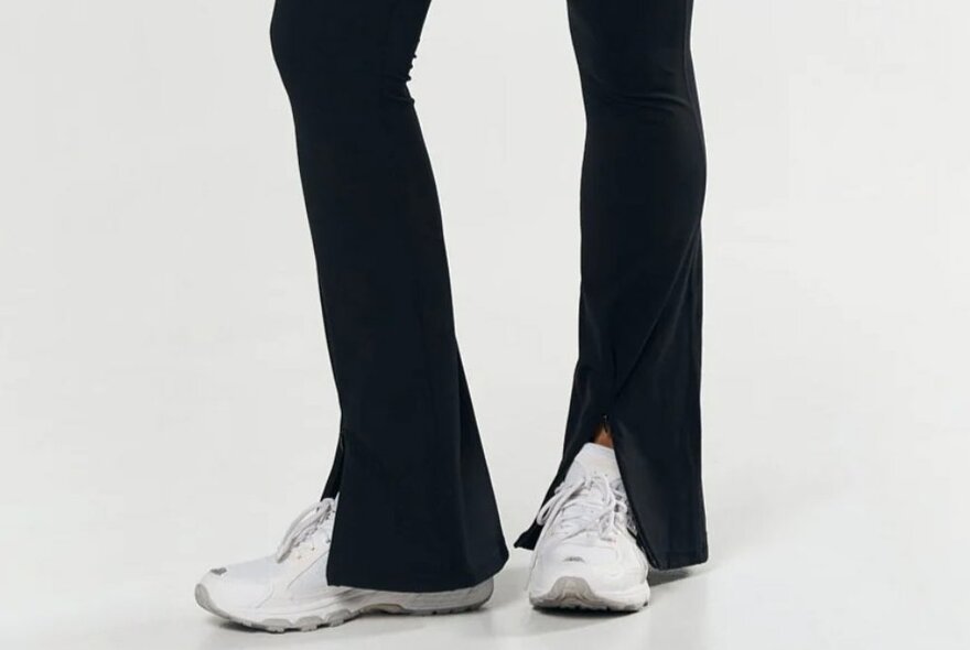 A model wearing black flared leggings with a split up the front showing white sneakers.