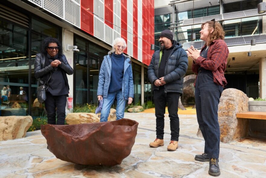 Four standing people smiling and interacting with each other in front of a low bronze sculpture in the courtyard at narrm ngarrgu Library.