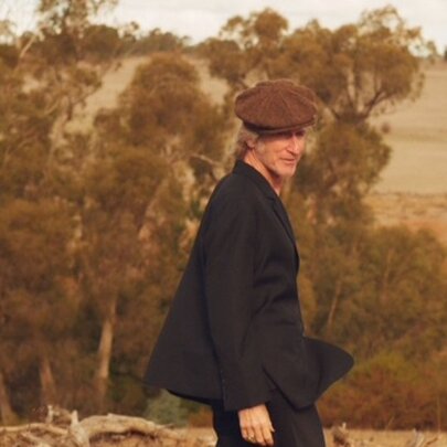 Musician and actor Tim Rogers wearing a dark suit jacket and brown cap, walking outside in a dusk tinged Australian bush landscape.