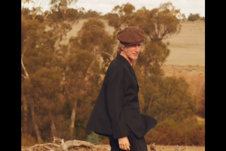 Musician and actor Tim Rogers wearing a dark suit jacket and brown cap, walking outside in a dusk tinged Australian bush landscape.