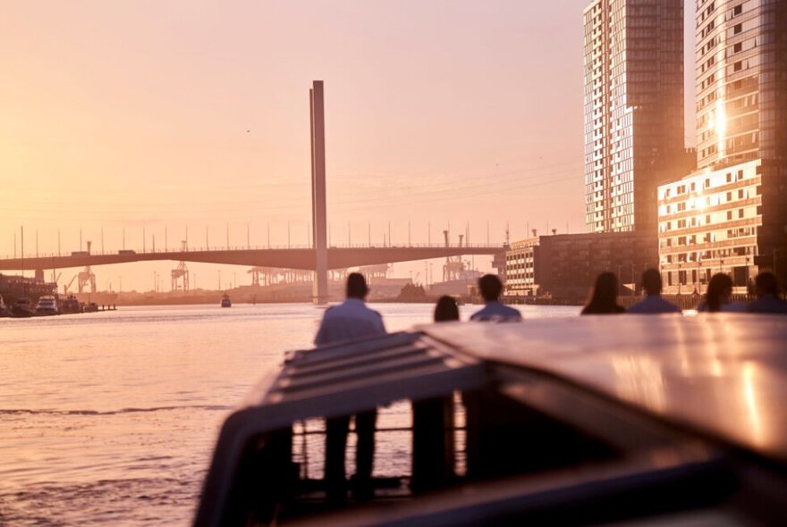 A view of Melbourne's Bolte Bridge at dusk from the top of a river cruise boat, with people standing in the front of the boat looking at the scenery.