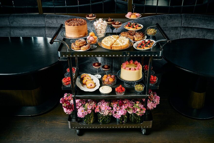 A three level dessert trolley filled with sweet treats and cakes, in front of booth seating inside a restaurant.