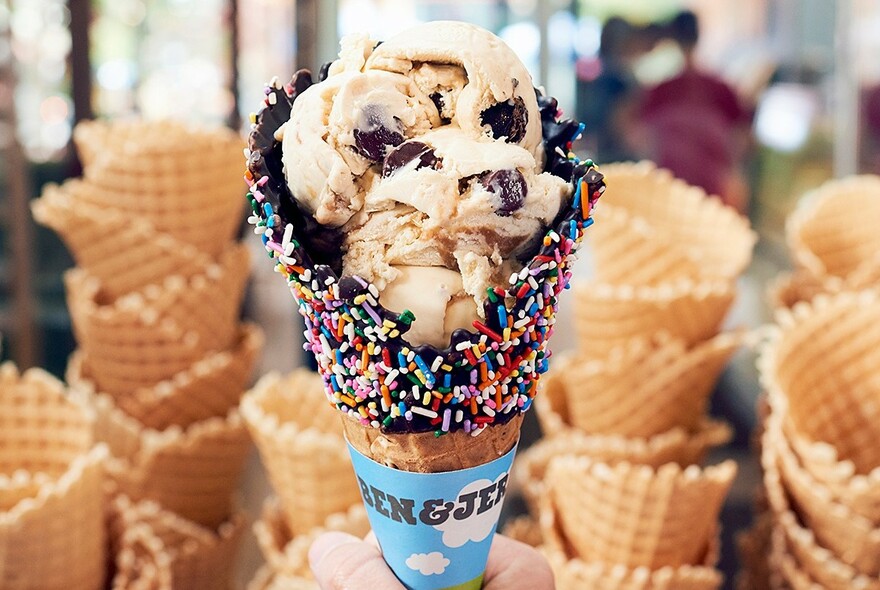 Icecream in a cone that's been choc-dipped and rolled in coloured sprinkles. Many cones in the background.