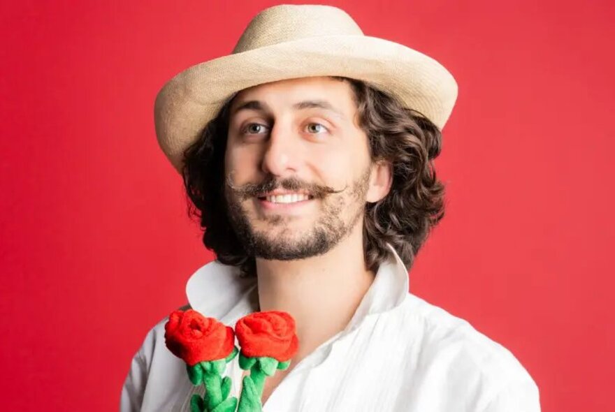 Jeromaia Detto in character as Giuseppe for his show, wearing a straw hat, a white shirt and holding two red roses, standing against a red background.