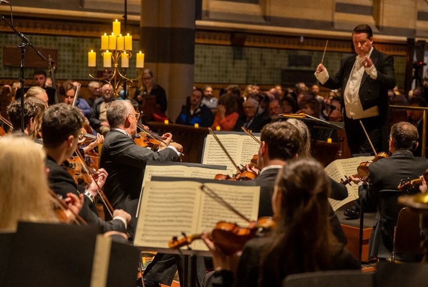 A conductor wearing a tuxedo, using a baton to conduct the orchestra in front of him, an audience seated behind him.