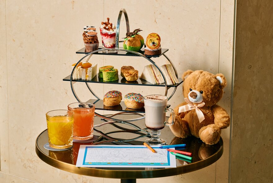 A children's high tea with a tiered stand of jungle-themed small food items, two glasses of juice, a colouring activity and a small teddy bear soft toy.