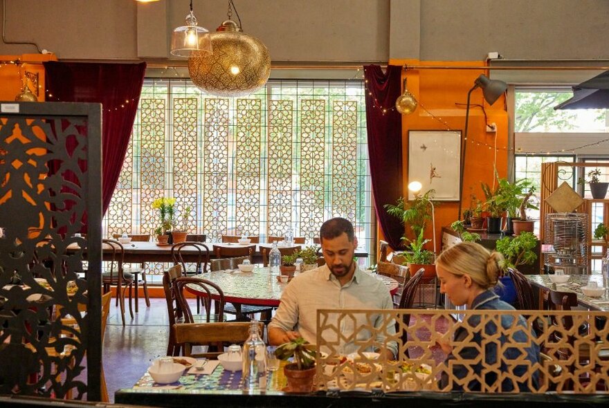 Restaurant interior showing a couple dining, plus other tables and chairs, set against a pattered street window, hanging light shades and orange painted walls.