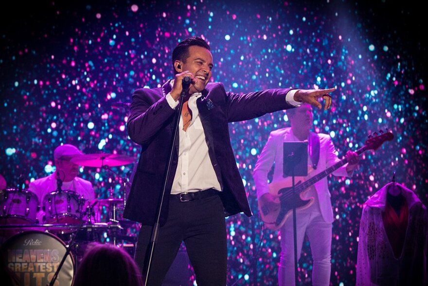 Singer in front of a band on stage, holding a microphone stand, pointing with his left arm and smiling.