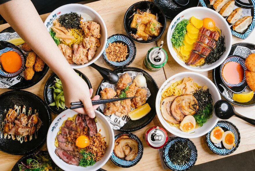 Japanese noodle dishes displayed on a wooden table, seen from above, with a hand picking up a piece of fried chicken with chopsticks.